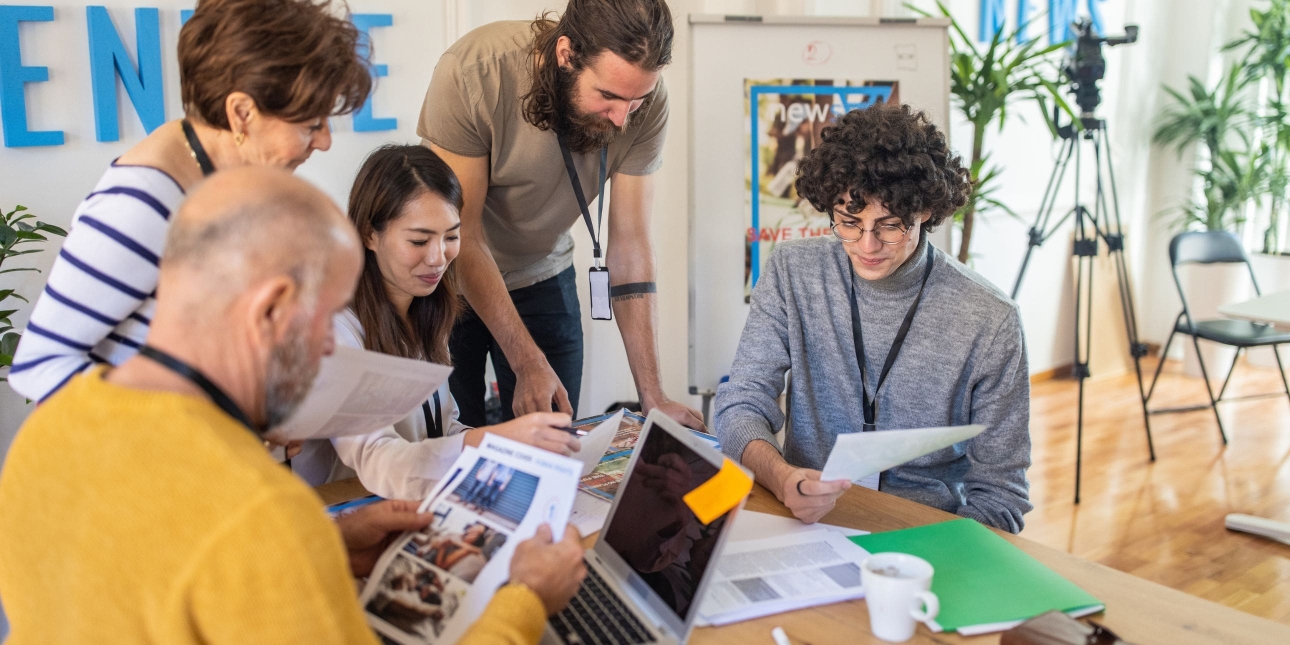Five casually dressed media agency colleagues of different genders and ethnicities gather around a desk examining papers and photographs. There is a camera on a tripod in the corner